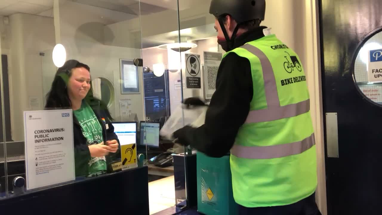A rider in a Chorlton Bike Deliveries vest handing a delivery through a health centre hatch